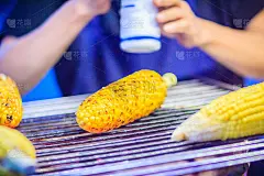 在烤制混合甜玉米黄油时，靠近街头食品摊贩的手。厨师正在烤，并在电炉上把盐、糖和黄油洒在烤好的甜玉米上
