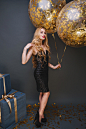 Indoor portrait of graceful curly lady posing with pleasure at her birthday party. glad female model in sparkle black dress standing near balloons and present boxes during event.