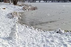 一个小雪人站在冰雪覆盖的森林里结冰的湖岸边。