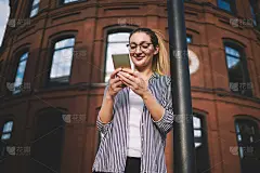 积极的嬉皮女孩在大型建筑附近等待朋友时，寻找现代电子产品的信息;20多岁的快乐女人在户外用高级智能手