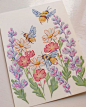 a card with watercolor flowers and bees on it, sitting on a white surface