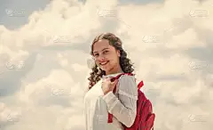 快乐的卷曲女孩背着背包在天空的背景下。回学校去童年的快乐阳光灿烂的日子里的小孩。暑假时间。小孩子在春