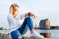 年轻美丽的微笑女孩穿着白衬衫和牛仔裤，像男孩子一样坐在栏杆上