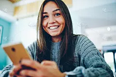 一个有着棕色头发的快乐年轻女子，在室内使用互联网连接安装现代智能手机上的新应用程序时，对着镜头微笑。