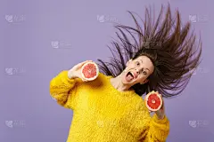 兴奋的年轻女子与流动的头发保持张嘴持有新鲜成熟的葡萄柚孤立在紫罗兰柔和的背景。人们生动的生活方式，放