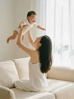 一位穿着白色吊带连衣裙的长发女子斜倚在柔软的米色沙发上，双手高高举起一个穿着白色连体衣的婴儿，婴儿悬空舒展着四肢，两人脸上洋溢着幸福灿烂的笑容。温暖柔和的自然光从窗外洒落，透过轻盈的白色纱帘形成朦胧的光影效果，照亮了整个室内空间。整体画面呈现出温馨、治愈的氛围，采用写实的摄影风格，色彩以柔和的米白色调为主，光影过渡自然，细节清晰，捕捉到母子间亲密互动的美好瞬间。