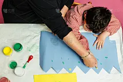 特殊教育的老师和学生穿着长袍，在桌子上用手画着生日花环和涂着丙烯颜料的黄色纸板。塑料工艺疗法。