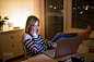 Woman at desk, holding smartphone, working on laptop at night.