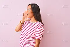 年轻的中国妇女穿着条纹t恤和眼镜在孤立的粉红色背景大声喊叫和尖叫，用手捂着嘴。通信的概念。