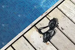 一个温暖的夏日，游泳镜放在一个木制的表面上，背景是一个模糊的游泳池。