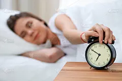 昏昏欲睡的年轻女子伸出手来敲响警钟，愿意关掉它。早醒、睡眠不足的概念