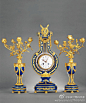 法国蓝珐琅瓷里拉琴式三件套钟，由一座拉琴式钟与两个花台组成。钟和花台均由铜质框架和蓝珐琅瓷里结合制成。钟为拉琴式，钟座为圆形，下承四个圆形铜质小支脚。瓷胎表面镶嵌铜质花卉纹与连珠纹。白珐琅瓷、水晶、黄金、蓝珐琅瓷，整体质感奢华高贵，尺寸：41厘米，钟：52厘米，成交价：RMB 1,150,000
