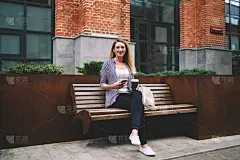 肖像的年轻积极的女人看相机，而等待朋友在长凳上与手机和咖啡在手里，快乐的潮人女孩穿着时髦的服装拿着现