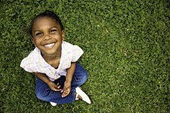 Portrait of adorable African American girlBeautiful preschool age African American little girl smiles while looking up at the camera. She is sitting in green grass.