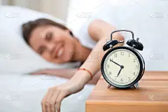 昏昏欲睡的年轻女子伸出手来敲响警钟，愿意关掉它。早醒、睡眠不足的概念
