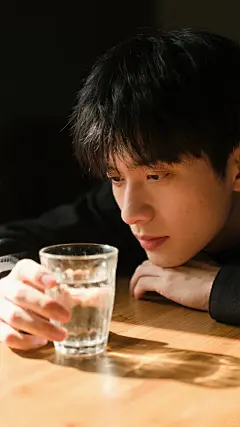 一位男性，黑色短发，神情专注，趴在木桌面凝视手中透明有水玻璃杯。光线从一侧洒落，形成明暗对比，增强层次感与氛围感。身着深色衣物，背景暗调，突出主体。构图特写，聚焦脸部与手部，自然光线柔和，色温偏暖，色彩饱和度适中，明暗过渡细腻，有明显噪点，质感高清。场景室内，光线营造静谧专注氛围，人物姿势低头凝视，手部轻柔触摸物体，传递沉静情感。整体画面光影艺术感强，展现摄影的质感与层次，给人视觉沉浸感。