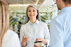 亚洲中年微笑女商人首席执行官经理谈话教年轻的白人男性和女性实习生同事讨论业务项目站在现代公司的办公室