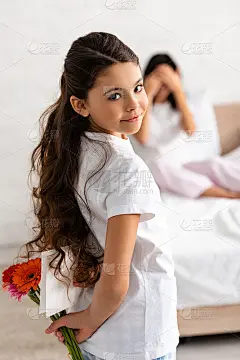 有选择的焦点可爱的孩子拿着鲜花和母亲节贺卡，而妈妈坐在床上，用手捂着眼睛