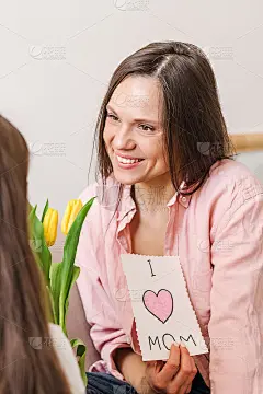 快乐的微笑黑发年轻女子接受她的女儿在母亲节的祝贺，并持有自制的贺卡。母亲节、3月8日和生日的概念