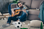Photo of hipster guy with long beard sitting carpet near sofa holding guitar don't mind chaos after stag party messy dirty flat singing songs careless person indoors
