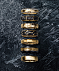 A color photograph shows a vertical row of seven David Yurman men’s rings standing upright on a scratched, dark grey icy surface dusted with snowflakes. The jewelry is crafted from 18K yellow gold. From top is a Beveled band with pavé white diamonds, a Fa