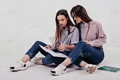 坐在地板上，阅读记事本上的一些信息。一对双胞胎姐妹坐在工作室里摆姿势，背景是白色