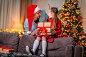 loving dad and daughter hug at home, sitting on couch against background Christmas tree. child and woman are laughing happily, holding gift box in their hands. parent gives little girl gift.