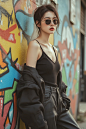 OBbllai, A stylish outdoor photograph of a model with a chic haircut and sunglasses, wearing a trendy outfit, confidently posing against a backdrop of colorful street art in a vibrant urban setting. The graffiti art adds a bold and edgy element to the sce