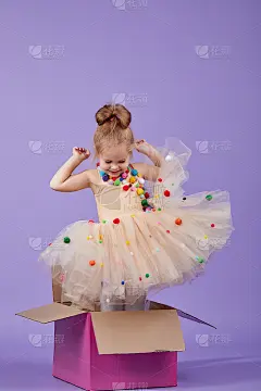 一个快乐的小女孩穿着华丽的节日服装坐在一个大的彩色礼品盒里，在紫色的孤立的背景上。惊喜的生日派对。空