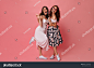 Brunette lady shows peace sign and poses with her friend on pink background. Girls with wavy hair in white fashionable dresses and converse hugging