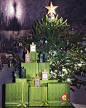In front of a black and white painted winter mountain scene, a festively decorated pine tree with a large gold star on top, stands behind a pyramid of green damask print Gucci gift boxes stacked on a table. Balanced on top of the boxes are bottles of thre