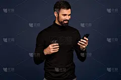 微笑的年轻人在时尚的毛衣摆姿势孤立在蓝色墙壁背景工作室肖像。看手机的人，拿着纸咖啡杯或茶