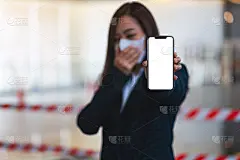 一名妇女戴着防护口罩，站在防控新冠肺炎概念传播的红白胶带前，展示白色手机屏幕