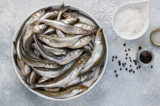 胡瓜鱼。在灰色的混凝土背景下，白色的碗里装着新鲜的生小鱼。香料-粗海盐和黑胡椒。选择性聚焦，俯视图