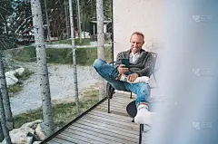 微笑的中年男子穿着开襟羊毛衫，牛仔裤和温暖的袜子坐在森林小屋的阳台上，使用着现代智能手机。日常生活照