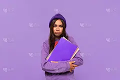 沮丧的非洲裔美国少年在一顶帽子和毛衣拿着文件夹孤立地站在紫色的背景。教育的概念