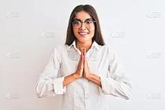 年轻美丽的女商人戴着眼镜，站在孤立的白色背景与双手一起祈祷，请求原谅自信的微笑。