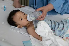 母亲抱着奶瓶给11个月大的男婴喂奶，男婴在病床上喝奶。卫生保健医疗。