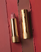 A still life features an open Gucci Rouge à Lèvres Satin – Satin Lipstick lying next its gold case with delicate floral inscription. The background is four different streaks of red and pink lipstick to demonstrate the different shades in the collection.