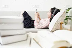 快乐的亚洲少女戴着耳机躺在沙发上，一边休息一边用手机上网听音乐，享受放松后的阅读，学习课本，放松概念