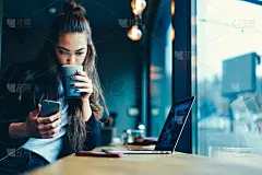 可爱的时髦女孩与长头发看有趣的视频，同时喝美味的饮料室内。一名年轻女学生在咖啡店用智能手机查看4G网