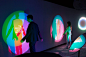 This may contain: two people standing in front of an art installation with colorful lights on the wall and behind them are silhouettes of people
