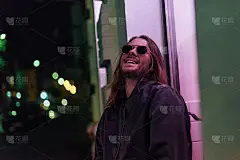 穿着皮夹克，戴着墨镜，笑着的年轻人在粉色的灯光下走在大街上