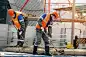 两名戴着安全帽、工作服和医用口罩的工人正在一个建筑工地用铁锹工作。重体力劳动