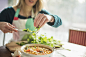 Mint Images在 500px 上的照片A woman wearing an apron, sitting at a table, sprinkling herbs into a bowl of vegetable stew.