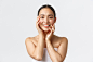 Gorgeous sensual asian woman in towel touching face and smiling, applying skincare products, cosmetic procedure at spa salon, massaging face and gazing camera happy, white background. Free Photo
