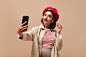 Stylish woman in red beret takes selfie on beige background. Beautiful lady with bright lipstick with earrings and in raincoat makes photo.