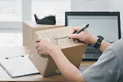 近手一名男子用钢笔在包裹盒上写下包裹的收货地址，并使用笔记本电脑在网上系统中注册
