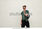 Stylish young handsome man with sunglasses in beach colored shirt with bag posing near green wall