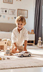 in a bright children's room,A real photo with a full-body shot of a 3-year- old boy playing Play with toy cars,on the carpet in the living room with toys around. She is wearing a light beige home outfit with beautiful facial featuresfain skin, brown hair,